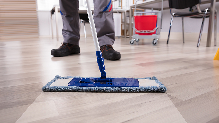 Bodenreinigung mit Wischmop Reinigungskraft wischt dem Boden mit einem feuchten Wischer. Im Hintergrund sind der Wassereimer, Stühle und Tische sowie ein Bücherregal zu sehen.